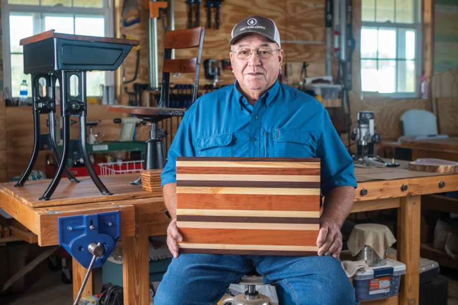 Randall Dasher holds a cutting board that he made from different types of wood, while seated in front of a historic school desk that he is refurbishing
